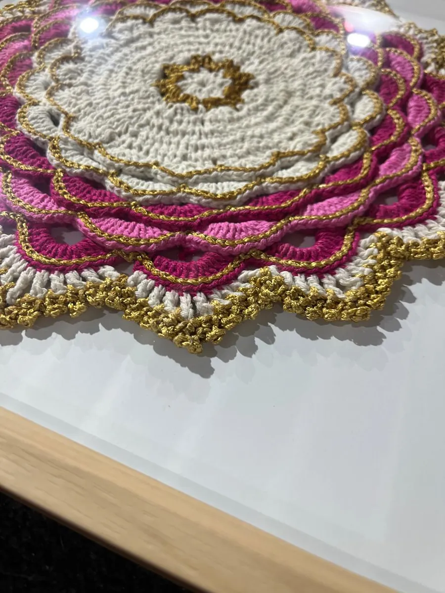 Mandala de crochê rosa, branca e dourada emoldurada em moldura de madeira clara com vidro